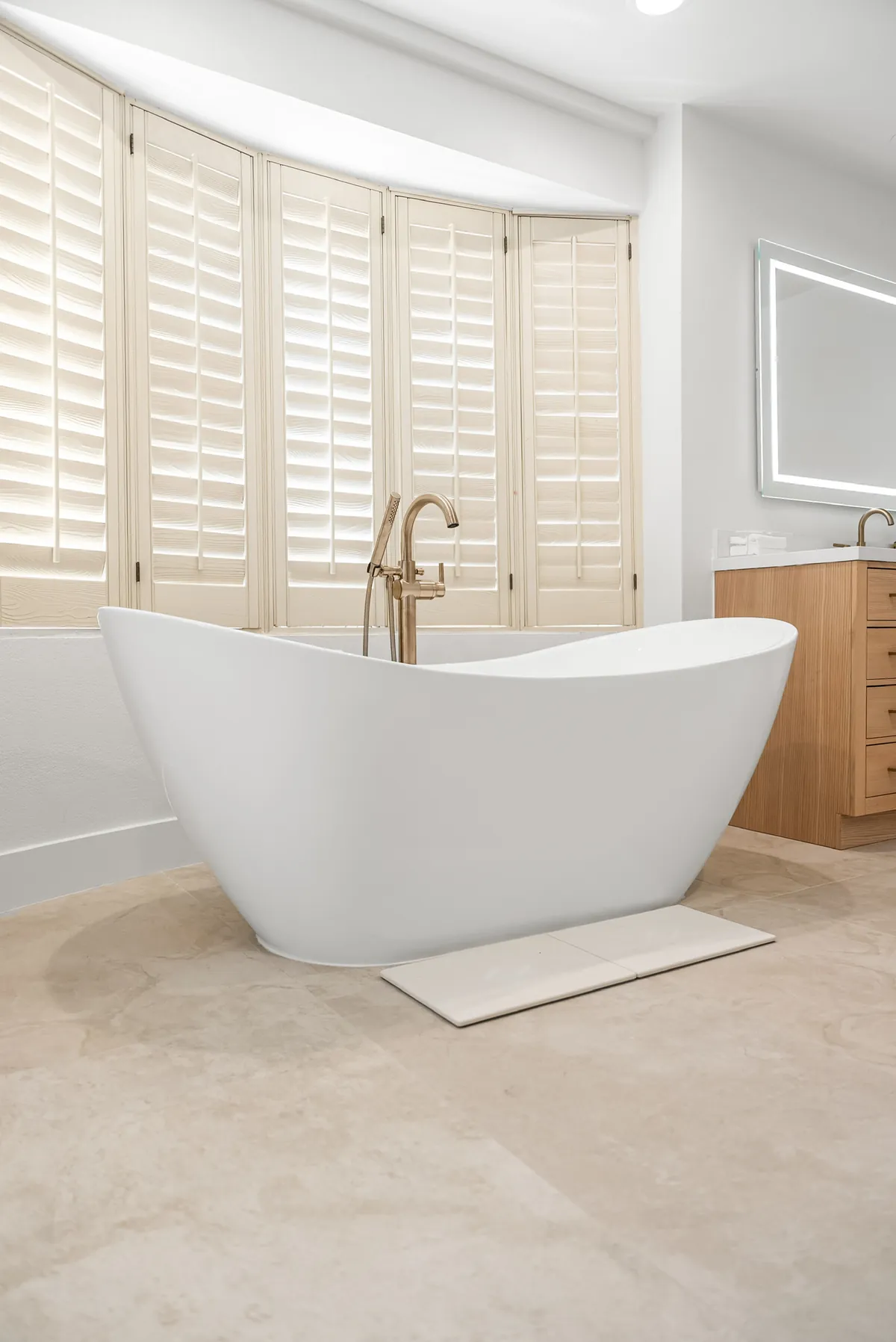 Freestanding tub under a bay of plantation shutters, with the vanity beyond